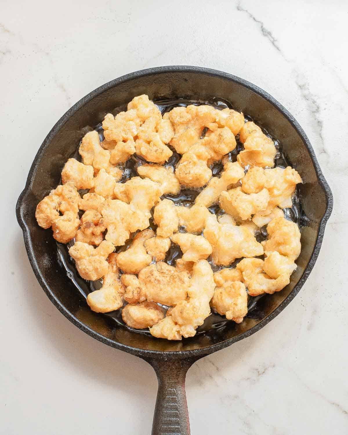 Golden fried cauliflower pieces sizzling in a cast iron skillet on a white marble surface.