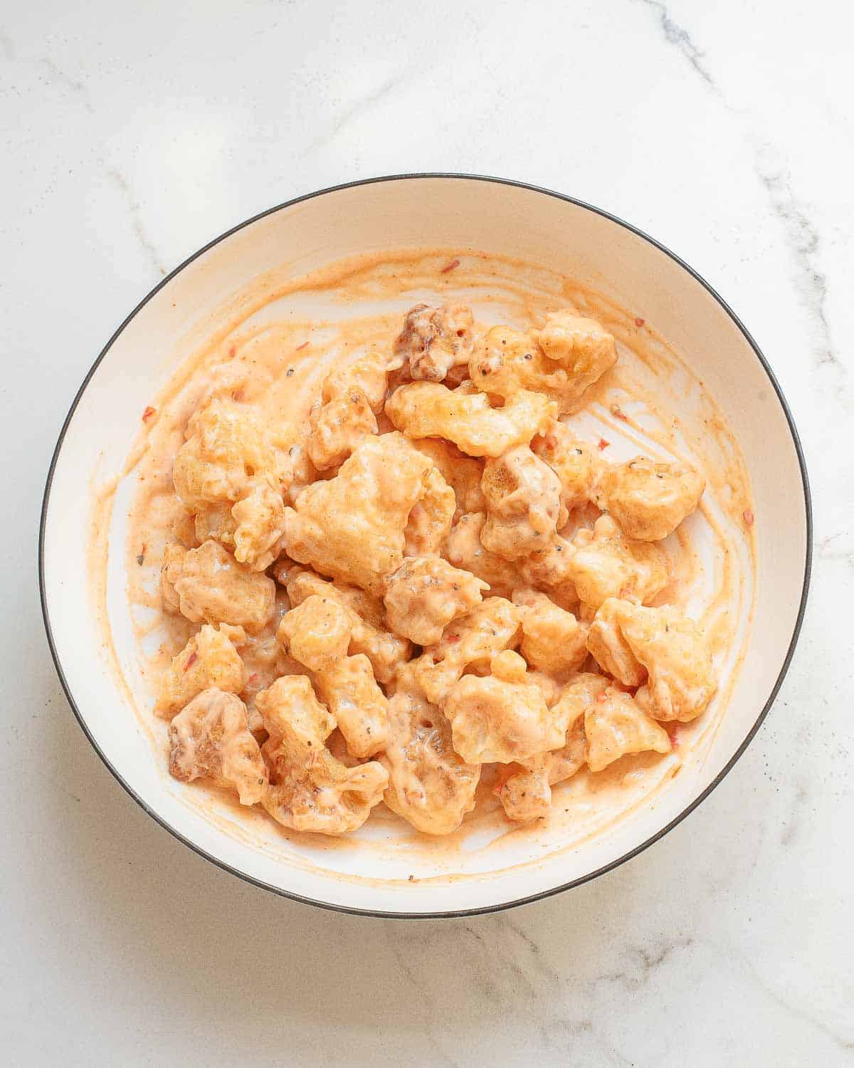 A bowl of pan-fried cauliflower pieces, coated with bang bang sauce, placed on a white, marbled surface.