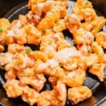 A black skillet contains golden, crispy cauliflower bites drizzled with a creamy bang bang sauce. Garnished with finely chopped green herbs for a pop of color, the dish has a deliciously inviting appearance.