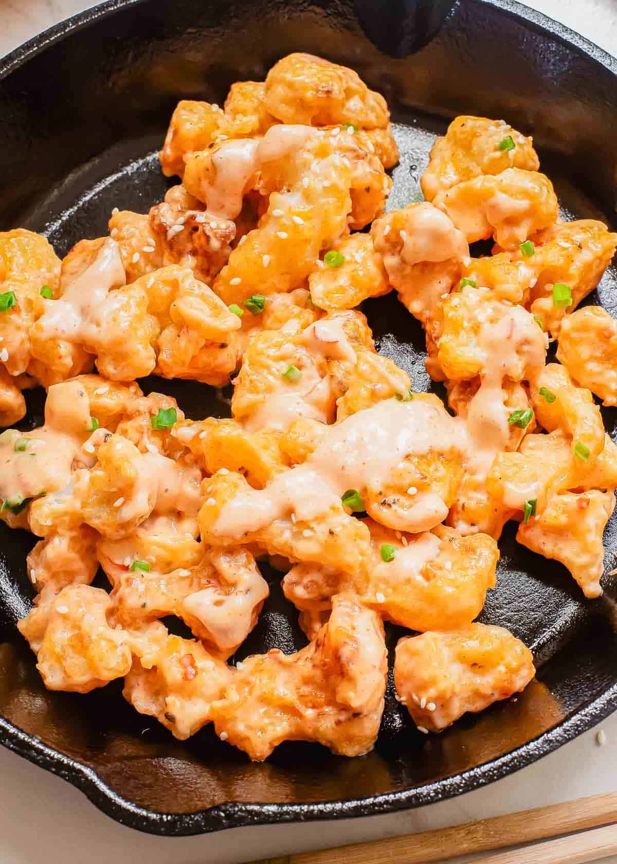 A black skillet contains golden, crispy cauliflower bites drizzled with a creamy bang bang sauce. Garnished with finely chopped green herbs for a pop of color, the dish has a deliciously inviting appearance.