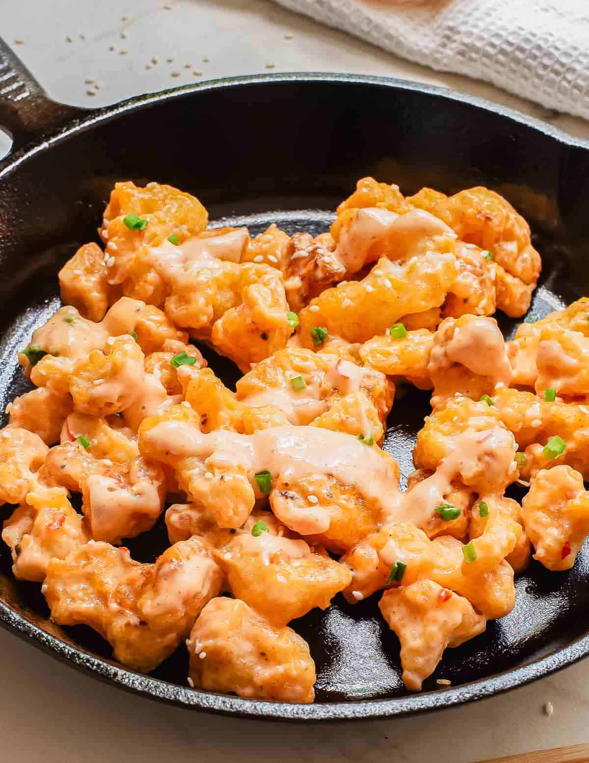 A close-up of a skillet filled with crispy, fried cauliflower bites drizzled with a creamy orange bang bang sauce, garnished with chopped green onions. The skillet sits on a light surface with a white cloth nearby.