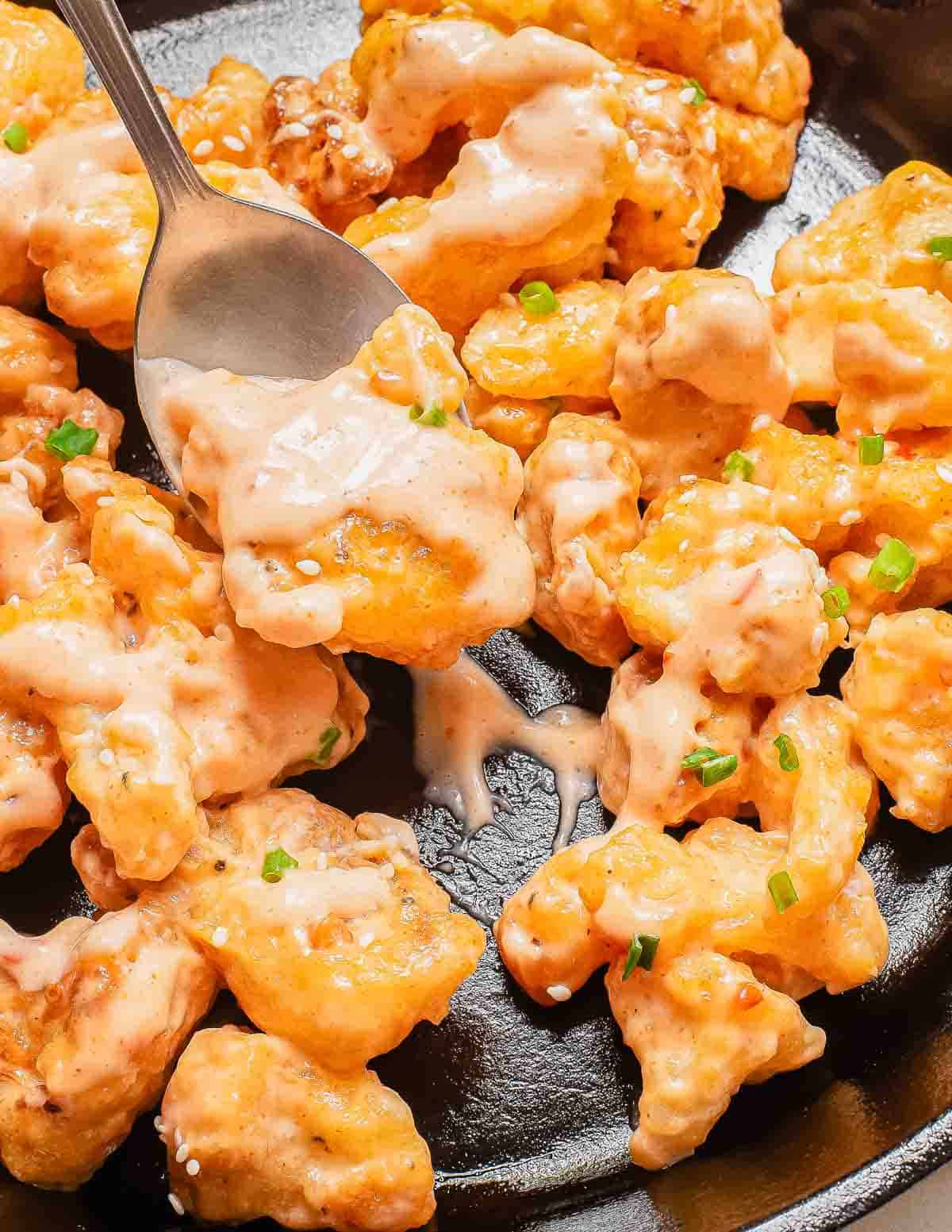 A spoon scoops up creamy, orange-hued battered cauliflower bites garnished with chopped green onions, served on a black skillet.