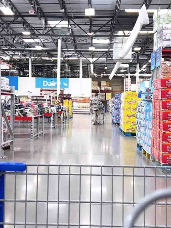 Inside a warehouse-style store, a shopping cart is pushed down an aisle stocked with large packages of snacks and drinks. The ceiling is high with exposed beams, and fluorescent lights illuminate the shelves. Shoppers are visible in the distance.
