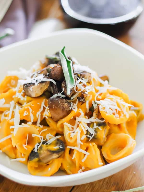 A bowl of creamy pasta topped with sliced mushrooms, grated cheese, and a sage garnish. The pasta is coated in a creamy butternut sauce and served in a white bowl on a wooden table.