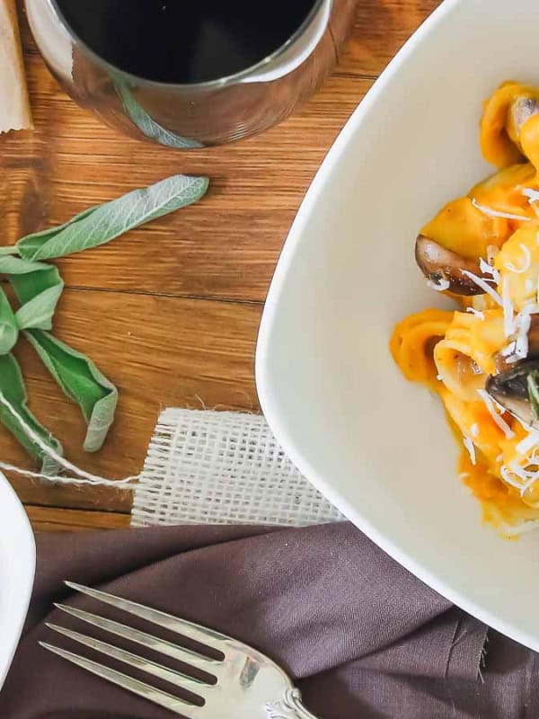 Two white bowls of butternut squash pasta with mushrooms, covered in grated cheese, are on a wooden table. A sprig of sage decorates one bowl. A glass of wine, a fork, and a small pile of cheese are nearby.
