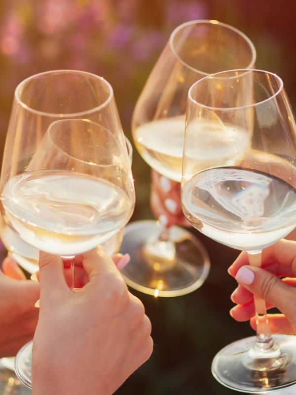 Four people clinking glasses of white wine outdoors in a celebratory manner, with a warm sunset in the background.