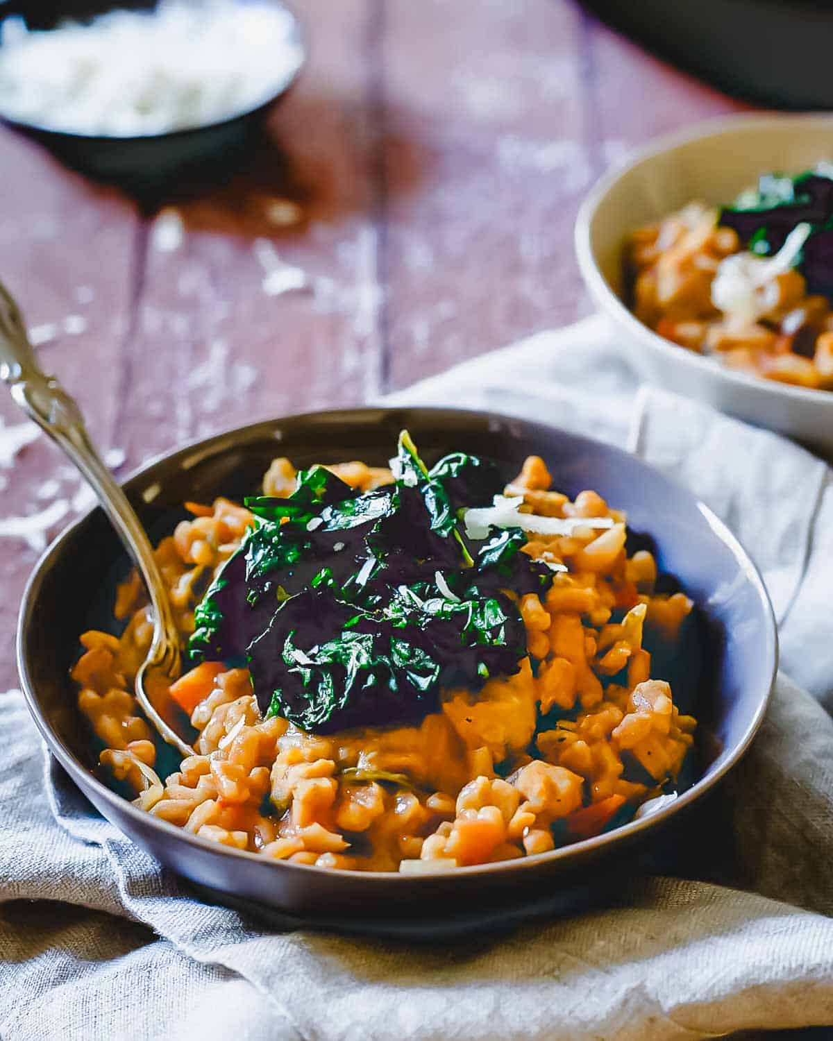 A bowl of creamy risotto topped with dark sautéed greens, and accompanied by a spoon. The risotto is on a wooden table with a cloth napkin underneath, and a second bowl and a dish of grated cheese are in the background.