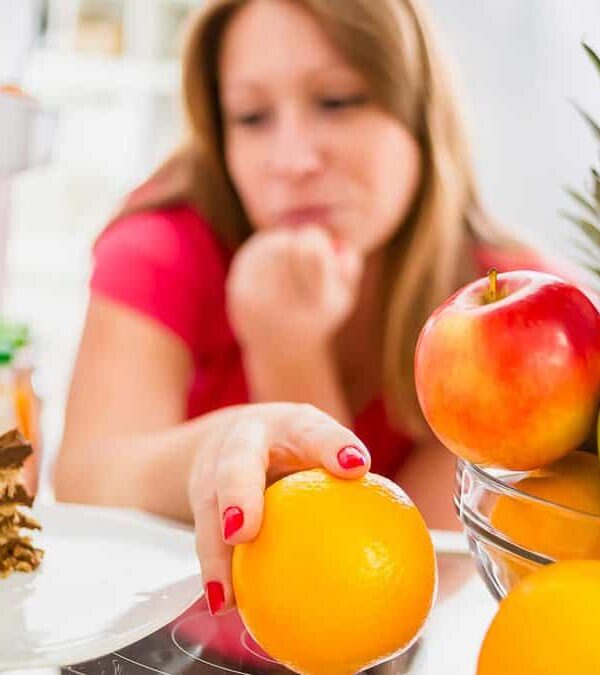 A woman in a red shirt reaches for an orange from a bowl of fruit inside an open refrigerator, while a slice of chocolate cake sits on a separate plate.