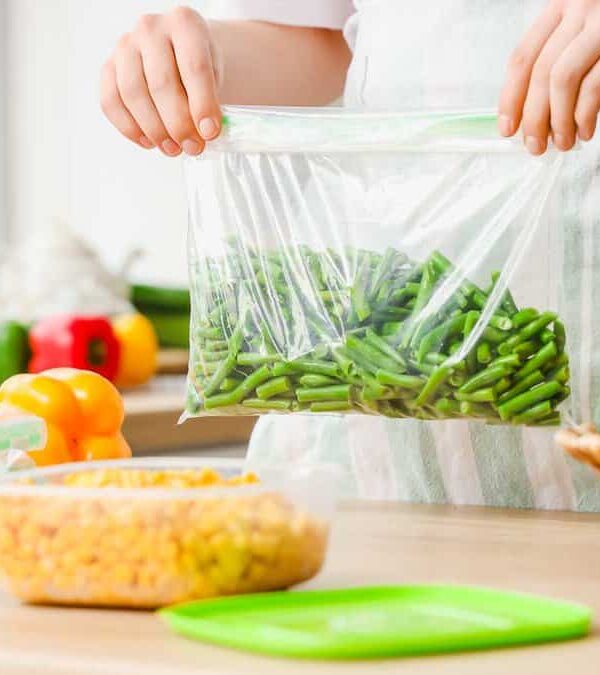 A person in a kitchen holds a clear plastic bag of green beans. The countertop displays various vegetables, including carrots, peas, corn, yellow peppers, and broccoli. The scene is bright and well-organized, suggesting meal prep.