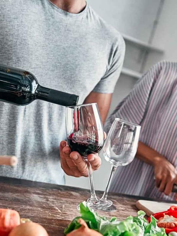 A man pours red wine into a glass while a woman smiles as she slices a red bell pepper. They are in a kitchen with various fresh vegetables on the counter, suggesting meal preparation.