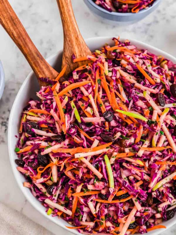 A colorful coleslaw with shredded red cabbage, carrots, and raisins in a white bowl, accompanied by wooden serving utensils. The vibrant salad is placed on a marble surface, with small bowls of additional slaw in the background.