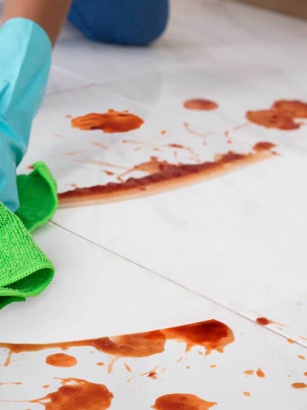 A person wearing blue gloves cleans up a red liquid spill on a white tile floor with a green cloth.