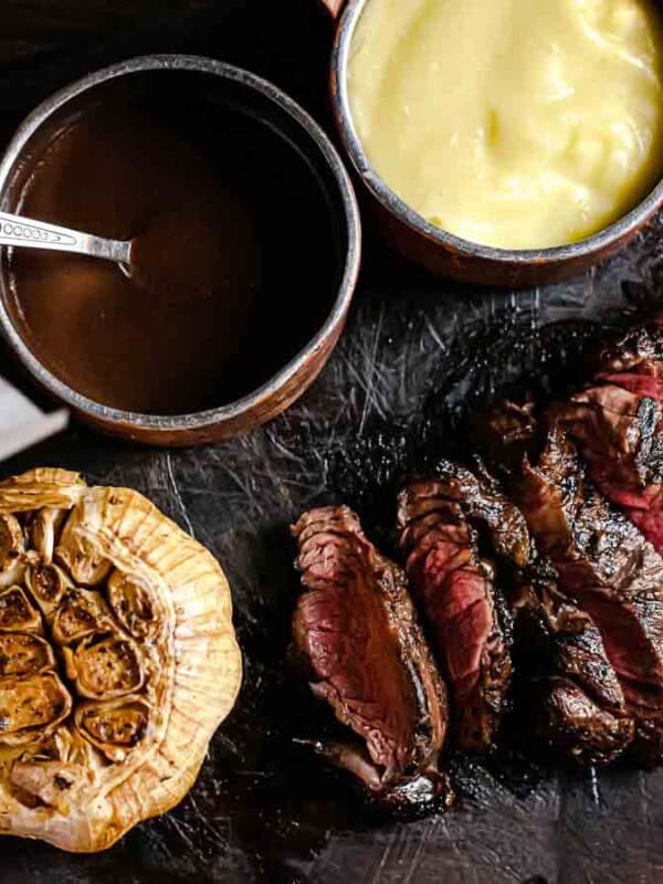 Grilled steak slices on a black slate board with roasted garlic, a copper bowl of gravy, a bowl of creamy sauce, and wooden containers with salt and pepper. A side of seasoned potatoes in paper is on the left.