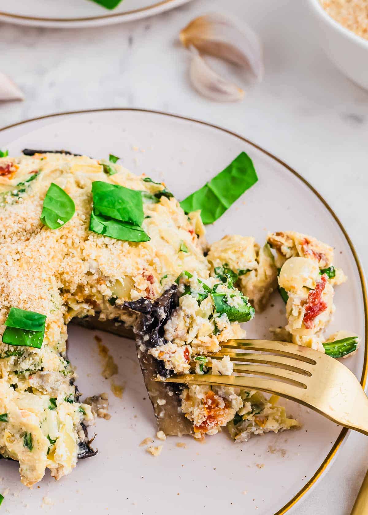 A close-up of a cheesy stuffed portobello mushroom on a white plate. It's garnished with chopped spinach and breadcrumbs. A fork is lifting a bite from the mushroom, with garlic cloves in the background.
