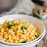 A bowl of sweet potato macaroni and cheese garnished with fresh green herbs. The dish is served in a white bowl on a rustic wooden table, accompanied by a fork and knife on a white napkin.