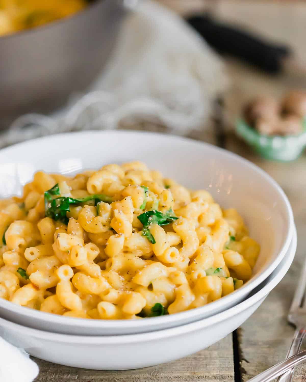 A bowl of sweet potato macaroni and cheese garnished with fresh green herbs. The dish is served in a white bowl on a rustic wooden table, accompanied by a fork and knife on a white napkin.