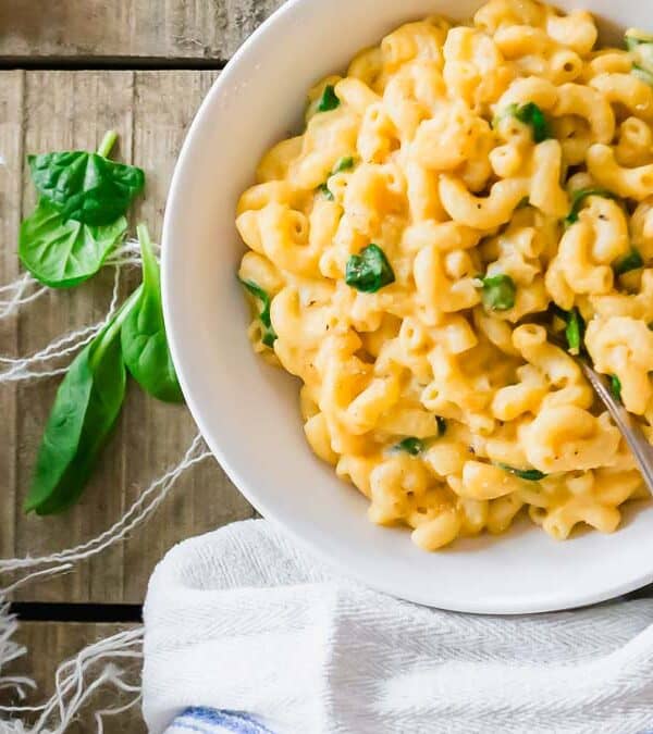 A bowl of macaroni and cheese made with creamy sweet potato sauce with baby spinach on a wooden table. A fork rests in the bowl, and a striped cloth is nearby. A pan with more pasta and spinach leaves is visible beside the bowl.