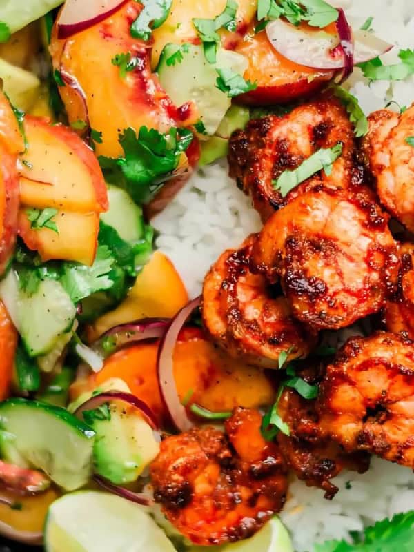 A vibrant dish featuring grilled shrimp on a bed of white rice, accompanied by a fresh salad of sliced peaches, avocado, red onion, and cilantro. Lime wedges are placed on the side for garnish, all served in a dark bowl.