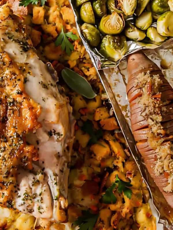 A top-down view of a roasted chicken breast with herbs, served on a bed of diced vegetables. Alongside are roasted Brussels sprouts and sliced sweet potatoes topped with crumbled nuts and herbs, all on a metal tray.