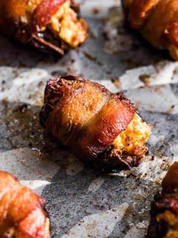 Close-up of stuffed dates wrapped in crispy bacon, placed on a baking sheet. The filling appears creamy and golden, contrasting with the dark dates and the browned bacon, showcasing a savory, appetizing snack.