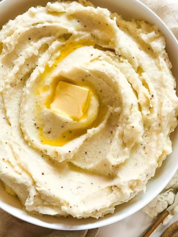 A bowl of creamy mashed potatoes topped with a pat of melting butter on a table. Surrounding the bowl are various vegetables and utensils, creating a rustic, inviting scene.