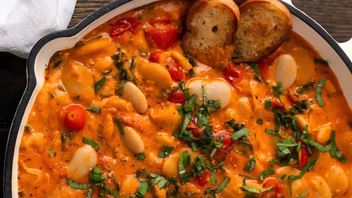 A close-up shot of a white skillet filled with a creamy tomato sauce with white beans, cherry tomatoes, and spinach. The skillet is rimmed with black, and there are two slices of toasted bread on the top of the sauce.