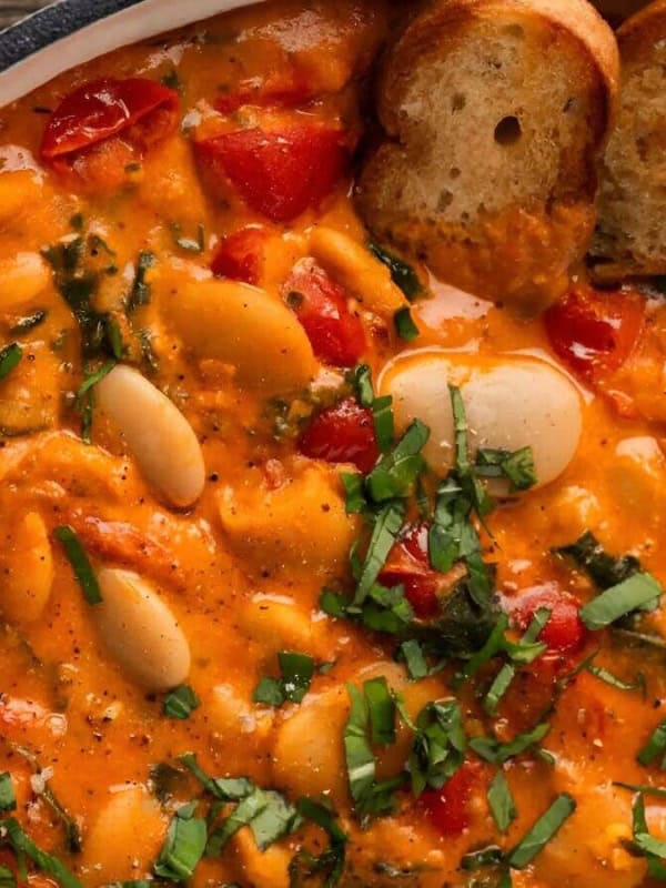 A close-up shot of a white skillet filled with a creamy tomato sauce with white beans, cherry tomatoes, and spinach. The skillet is rimmed with black, and there are two slices of toasted bread on the top of the sauce.
