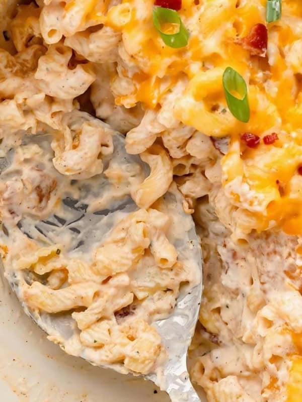A close-up of a creamy macaroni and cheese casserole in a baking dish. The dish is topped with melted cheddar cheese, bacon bits, and sliced green onions. A serving spoon is scooping a portion from the corner.