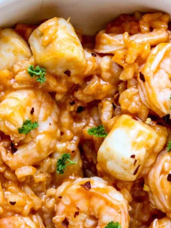 A bowl of creamy seafood risotto topped with shrimp, scallops, and garnished with fresh parsley. The dish has a rich, orange hue, suggesting the presence of a savory sauce, and is set against a light background.