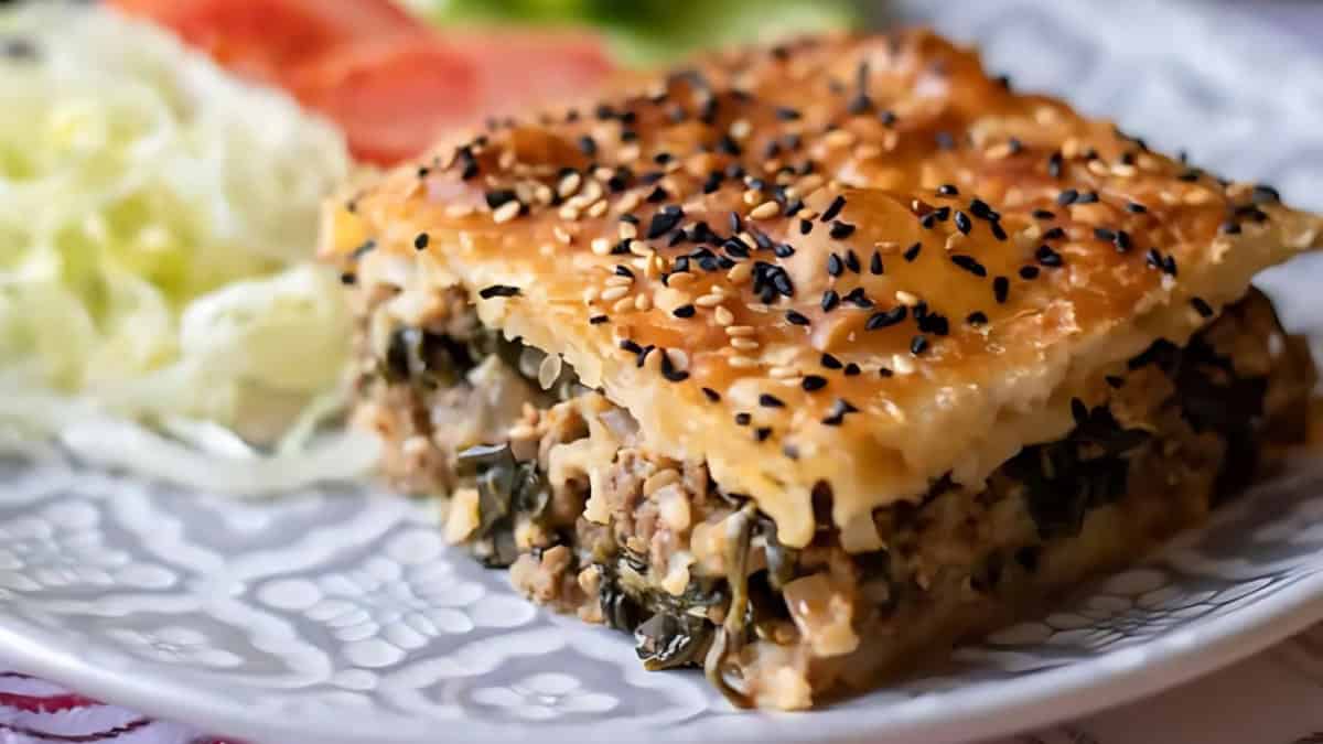 A close-up of a savory pastry, featuring layers of flaky, golden-brown dough topped with sesame seeds. The filling includes minced meat and greens. In the background, there are fresh salad ingredients on the plate.