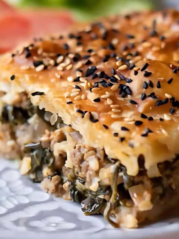 A close-up of a savory pastry, featuring layers of flaky, golden-brown dough topped with sesame seeds. The filling includes minced meat and greens. In the background, there are fresh salad ingredients on the plate.