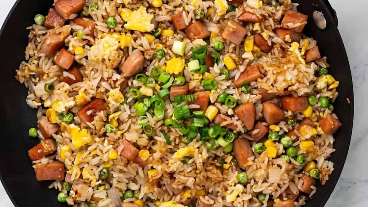 A skillet filled with fried rice mixed with diced ham, scrambled eggs, green peas, corn, and chopped green onions.