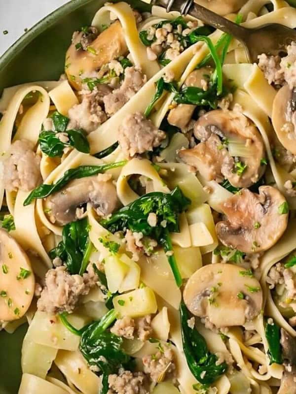 A plate of fettuccine pasta with sautéed mushrooms, spinach, and ground meat, garnished with chopped herbs. The dish is surrounded by additional plates of the same meal on a light background.