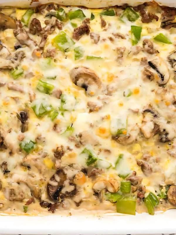A baked dish in a white casserole dish filled with melted cheese, ground meat, sliced mushrooms, and diced green bell peppers. The edges are slightly browned, and a checkered cloth is partially visible underneath.