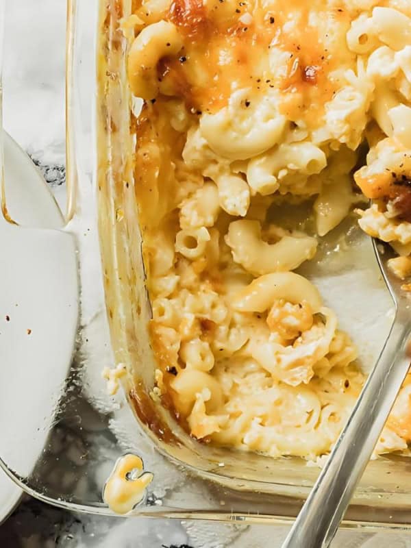 A glass baking dish filled with creamy macaroni and cheese topped with a golden crust. A portion is served on a white plate next to the dish, with visible melted cheese and black pepper sprinkled on top. A spoon rests inside the dish.