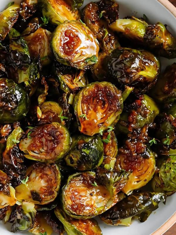 A bowl of roasted Brussels sprouts, caramelized and golden-brown, garnished with fresh thyme. The wooden table in the background adds a rustic touch to the presentation.