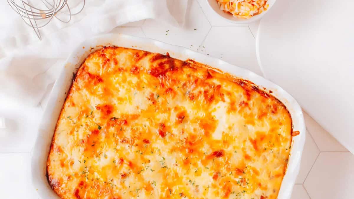 A freshly baked casserole with a golden, cheesy crust sits on a white counter. It is garnished with herbs. Nearby, a bowl contains grated cheese and kitchen utensils are visible. The bright setting highlights the casserole's appetizing appearance.