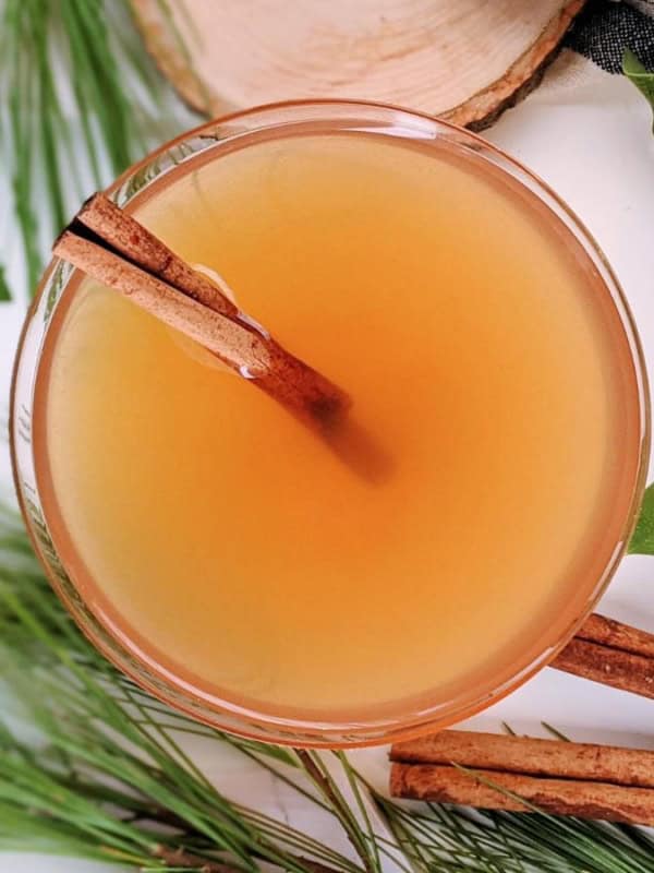 A glass of apple cider with two cinnamon sticks inside is surrounded by pine branches and green leaves on a white background.