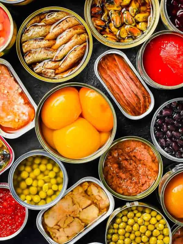 An assortment of opened canned foods arranged on a black surface, including vegetables, fruits, fish, and beans, showcasing a variety of colors and textures.