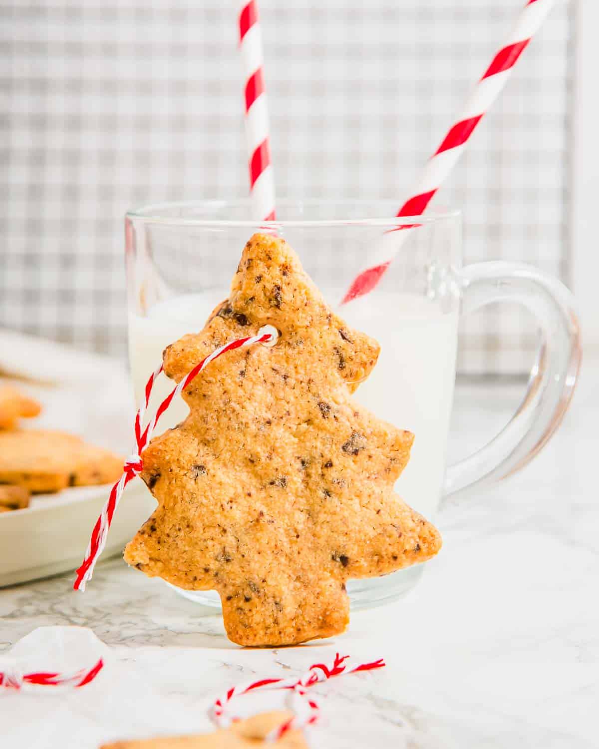A Christmas tree cookie standing up against a glass of milk with two straws in it.
