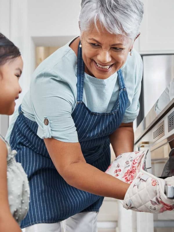 A smiling older woman wearing an apron uses an oven mitt to open the oven. Beside her, a young girl watches, also smiling. They are in a bright kitchen, sharing a joyful moment.