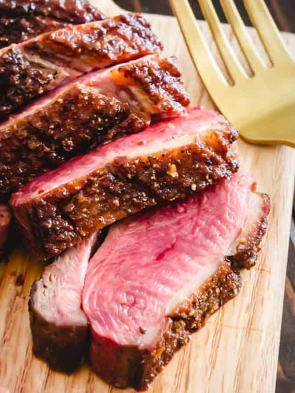 Sliced medium-rare steak on a wooden board with a small bowl of berry sauce beside it. A gold fork rests nearby, and a sprig of parsley is visible in the corner.