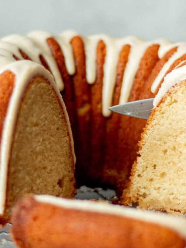 A freshly baked bundt cake with icing drizzle sits on a wire rack. A slice has been cut and placed in front, showing the cake's moist interior. A knife is poised near the cut section.