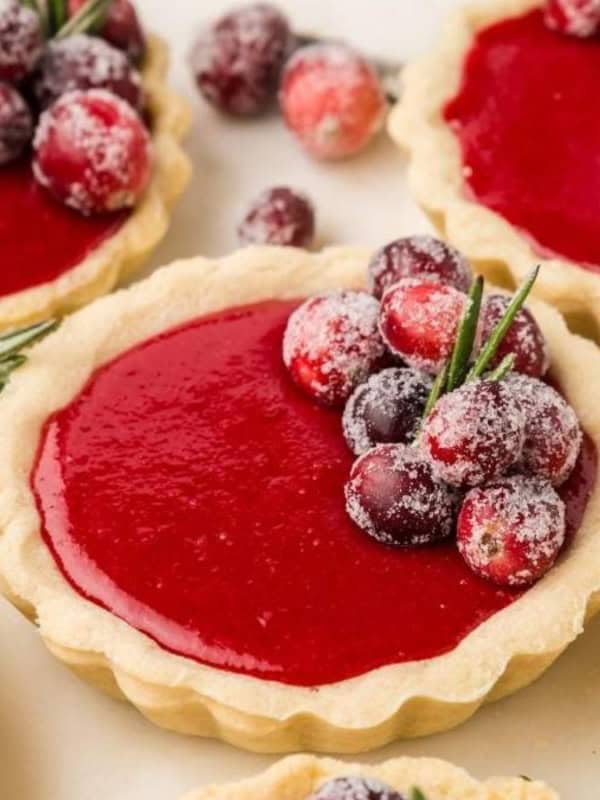 Mini tarts filled with vibrant red cranberry sauce are garnished with sugared cranberries and sprigs of rosemary. They are placed on a light surface, offering a festive and appetizing display.