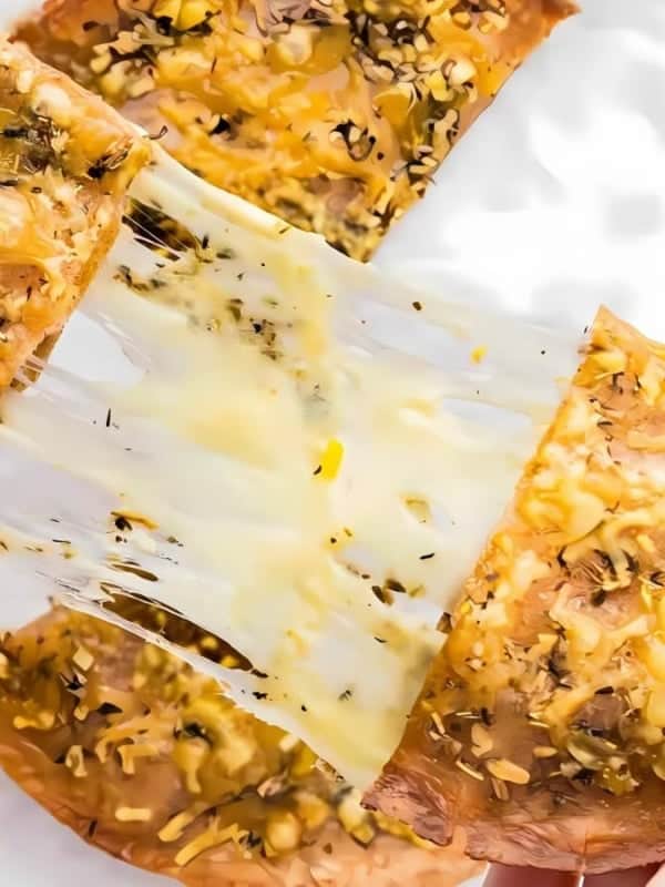 Two hands pull apart a cheesy, herbed flatbread, causing melted cheese to stretch. The flatbread is set on a white surface with more flatbread pieces visible in the background.
