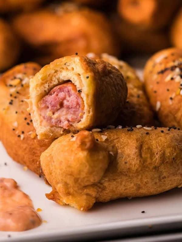A plate of golden-brown pigs in a blanket with poppy seeds and a side of pink dipping sauce. One is bitten to reveal the sausage inside. More snacks are blurred in the background.