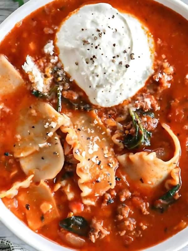 A bowl of lasagna soup filled with lasagna noodles, ground meat, spinach, and topped with ricotta cheese. The hearty tomato-based broth is garnished with black pepper. Fresh herbs and a napkin are visible in the background.
