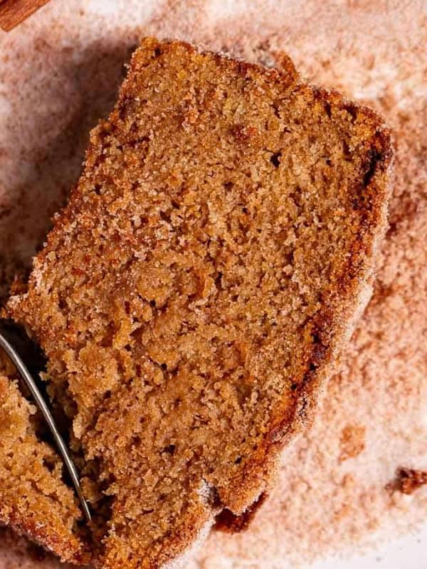 A close-up of a slice of brown, crumbly bread on a white plate dusted with cinnamon sugar. A fork is cutting through the slice, capturing the texture and spices. Another piece of bread is partially visible in the top right corner.