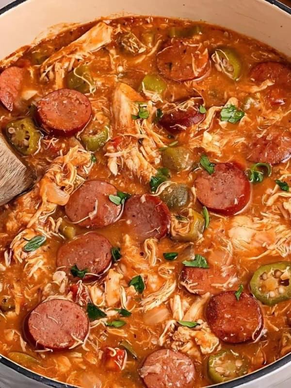 A pot of gumbo filled with sliced sausage, shredded chicken, and green bell peppers, simmering in a rich, reddish-brown broth. A wooden spoon rests inside the pot, surrounded by salt, pepper shakers, parsley, and bread on a kitchen counter.