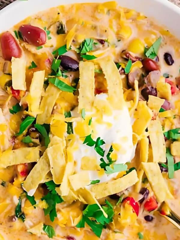 A bowl of creamy chicken tortilla soup garnished with shredded cheese, tortilla strips, chopped cilantro, and a dollop of sour cream. Beans and corn are visible in the soup. The bowl is resting on a marble surface.